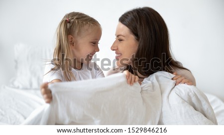 Happy young mother and little daughter making bed together, having fun, looking at each other profile close up, smiling mum and adorable preschool child holding blanket, relaxing in bedroom Royalty-Free Stock Photo #1529846216