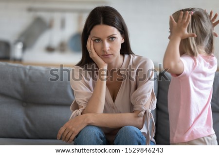 Unhappy mother having problem with noisy naughty little daughter screaming, demanding attention, child tantrum manipulation concept, tired thoughtful mum holding head, sitting on couch at home Royalty-Free Stock Photo #1529846243