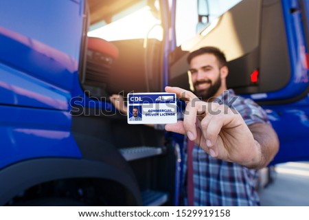 Portrait of middle aged bearded truck driver standing by the truck and showing his commercial driver license. Focus on CDL license. Truck driving school and job openings. Royalty-Free Stock Photo #1529919158