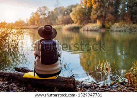 Tourist with backpack sitting on river bank at sunset. Middle-aged woman admiring autumn nature. Travelling concept