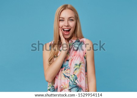 Joyful attractive blonde lady with long hair wearing natural make up, holding palm on her cheek and smiling widely to camera while posing over blue background
