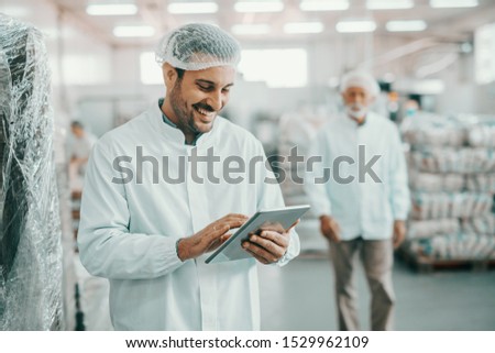 Smiling Caucasian supervisor in sterile uniform standing in food factory with tablet in hands and checking quality of goods.