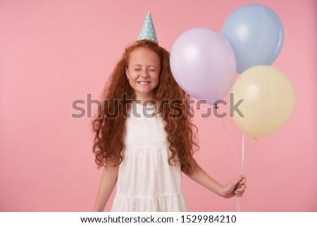 Studio photo of charming redhead female kid with long curly hair celebrates holiday, posing over pink background in festive clothes and birthday cap, smiling happily with closed eyes