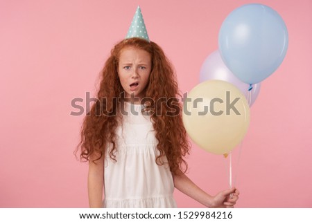 Studio shot of curly redhead female kid holding air balloons in hand while standing over pink background, wearing white dress and birthday cap, frowning and looking at camera with confused face