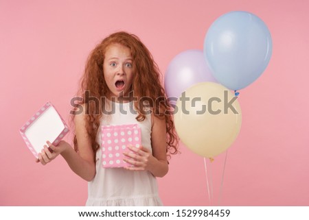 Excited lovely female curly kid with long foxy hair looking at camera with wide mouth opened, being surprised to get birthday present, standing over pink studio background in festive clothes