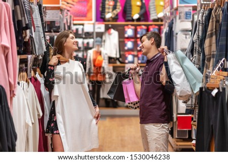 A smiling, happy girl tries on a dress that she really liked, and the guy looks at her approvingly, holding large bags with purchases in his hand. Emotions of joy and delight. Black Friday.