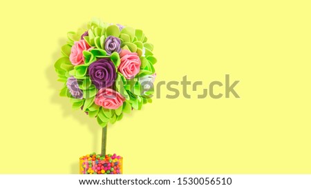 Bouquet of roses with green leaves handmade with foamy plastic with round shape planted in a glass pot with colored balls on yellow background with copy space