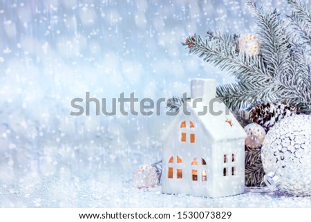 christmas decorations - toy glass house, balls and lights - near christmas tree branch under snow. white winter background