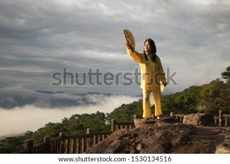 Asian woman practices Tai Chi on top of a mountain. Woman in traditional Chinese costume doing morning exercises with Chinese fans. 