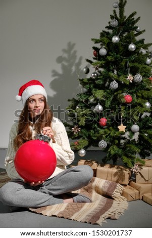 A young girl in a Christmas hat and a white knitted sweater with a large red ball in her hands sits on a plaid against the background of a Christmas tree. Christmas and new year concept.