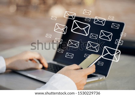 Side view of hands using laptop and smartphone on desk with digital emails on blurry office wall background. E-mail network and communication concept. Double exposure 