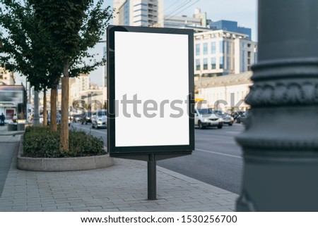 Vertical medium-size billboard with blank display for digital ads and promotional videos. Urban architecture in city center and pedestrian zone. Multistoried administrative buildings and living houses