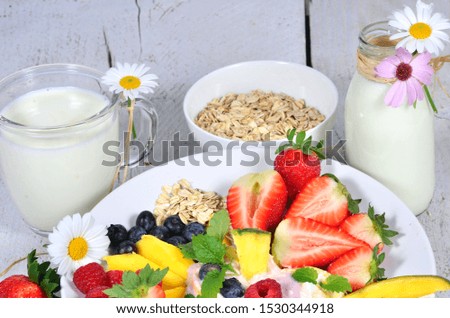 Enjoy your healthy breakfast: Close-up of a bottle and glass of milk, different fruits (strawberries, blueberries, pineapple, mangoes, raspberries) and oat flake decorated with white and pink daisies.