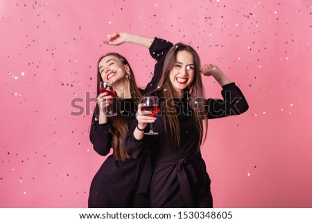Two women celebrate the New Year party having fun laughing under the flying confetti and drinking wine. Pink background, cheerful emotions, red lips, wearing black stylish evening outfits