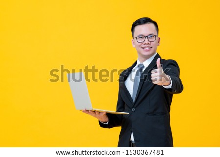 Young Asian businessman being happy with his online business carrying laptop computer and giving thumbs up isolated on yellow studio background