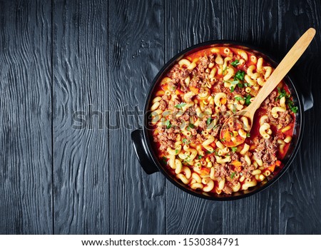 classic american goulash of elbow pasta, beef, celery and tomatoes in a black ceramic dish with a spoon, on a wooden table, view from above, flatlay, free space