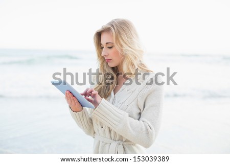 Pensive blonde woman in wool cardigan using a tablet pc on the beach