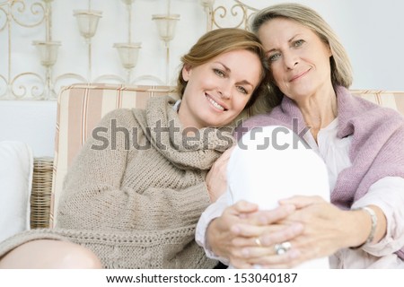 Close up family portrait of an adult daughter with her mature mother leaning on each other and being affectionate while relaxing on a sofa at home, outdoors.