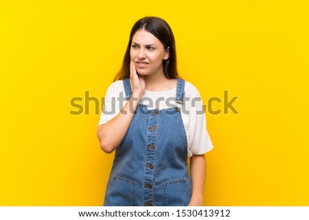 Young woman in dungarees over isolated yellow background with toothache