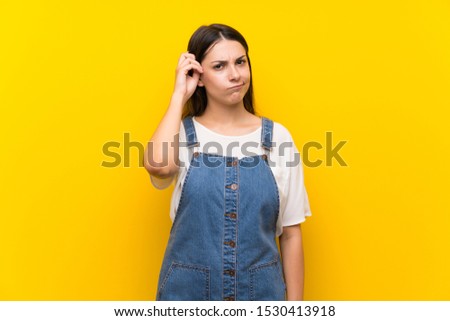 Young woman in dungarees over isolated yellow background having doubts