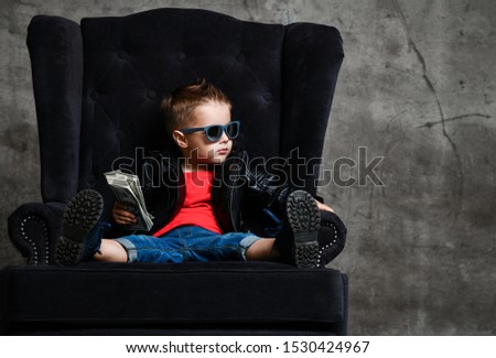 Self-confident rich kid boy in sunglasses, leather jacket, blue jeans and brutal shoes is sitting in big luxury armchair holding a bundle of dollars cash looking at free copy space