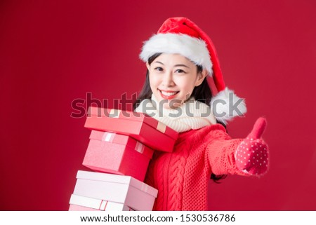 asian woman wear christmas hat and gloves holding presents and thumb up isolated on red background