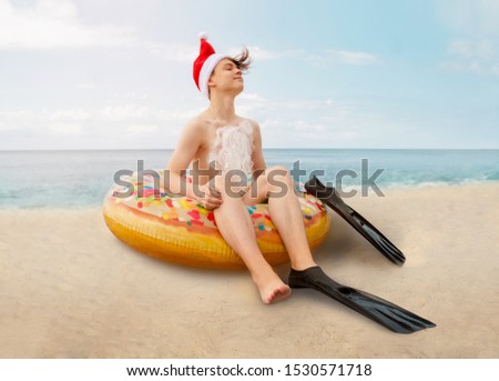 Teenage boy in flippers, Santa hat and inflatable circle resting on sea beach with blue sea and yellow sand