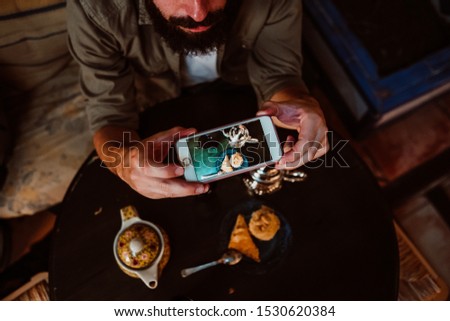 
Young handsome tourist enjoying a moment alone in a Moroccan style tea shop. Drinking traditional tea and eating typical Arabic sweets while taking pictures with his smartphone. Lifestyle
