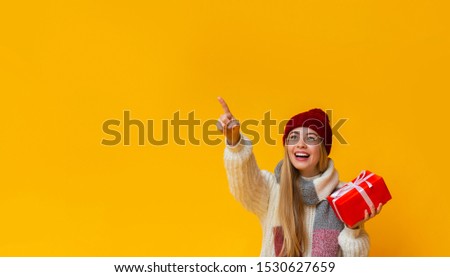 Happy girl in red winter hat holding Christmas present, pointing at empty space, yellow studio background