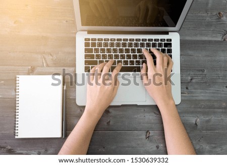 The hand of Asian woman typing on keyboard of laptop, maybe looking for information of education, check email of work or online shopping. On gray wood background with white notebook and pen, top view