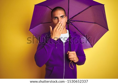 Young handsome man wearing purple umbrella over yellow isolated background cover mouth with hand shocked with shame for mistake, expression of fear, scared in silence, secret concept