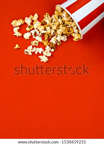 Spilled popcorn in a striped red and white cardboard box on a red background. Cinema, movies and entertainment concept. Top view, copy space.