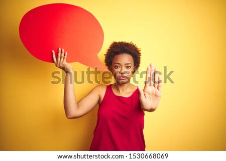 Young african american woman holding speech bubble over yellow isolated background with open hand doing stop sign with serious and confident expression, defense gesture