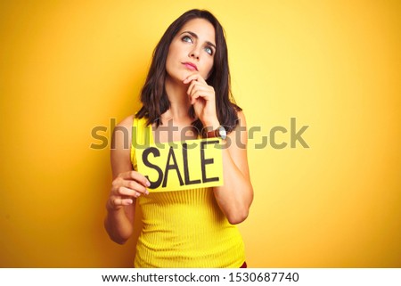 Young beautiful woman holding sale poster over yellow isolated background serious face thinking about question, very confused idea