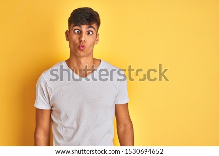 Young indian man wearing white t-shirt standing over isolated yellow background making fish face with lips, crazy and comical gesture. Funny expression.