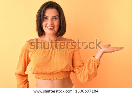 Young beautiful woman wearing casual t-shirt standing over isolated orange background smiling cheerful presenting and pointing with palm of hand looking at the camera.