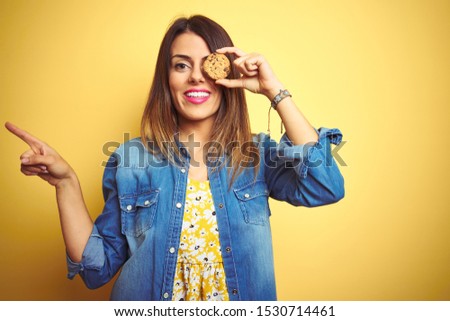 Young beautiful woman eating chocolate chips cookie over yellow background very happy pointing with hand and finger to the side