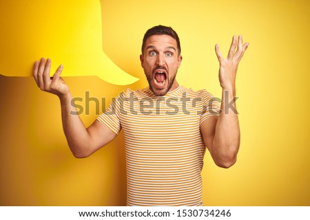 Young man holding empty speech bubble for message over yellow isolated background very happy and excited, winner expression celebrating victory screaming with big smile and raised hands