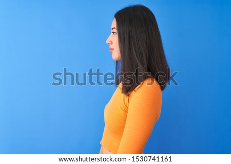 Young beautiful chinese woman wearing orange t-shirt standing over isolated blue background looking to side, relax profile pose with natural face with confident smile.
