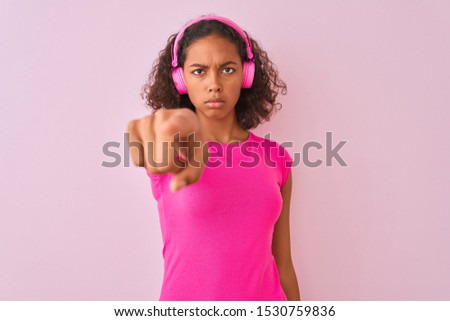 Young brazilian woman listening to music using headphones over isolated pink background pointing with finger to the camera and to you, hand sign, positive and confident gesture from the front
