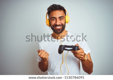 Arab indian gamer man playing video game using headphones over isolated white background screaming proud and celebrating victory and success very excited, cheering emotion