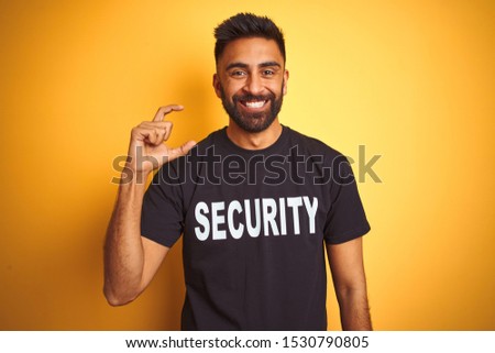 Arab indian hispanic safeguard man wearing security uniform over isolated yellow background smiling and confident gesturing with hand doing small size sign with fingers looking and the camera. Measure