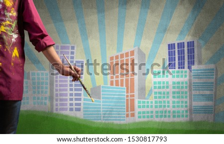 Close up artist hand holding paintbrush. Painter in shirt standing on background colorful picture of city. Modern downtown with high office buildings artwork. Creative hobby and artistic occupation.