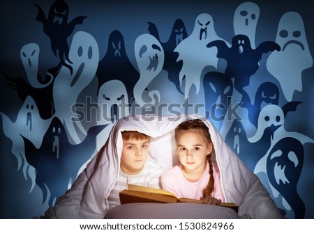 Cute girl and boy reading kids book in bed. Children with flashlight lying under blanket together. Kids in pajamas and funny ghosts silhouettes back on night sky. Children reading magic fairy tales