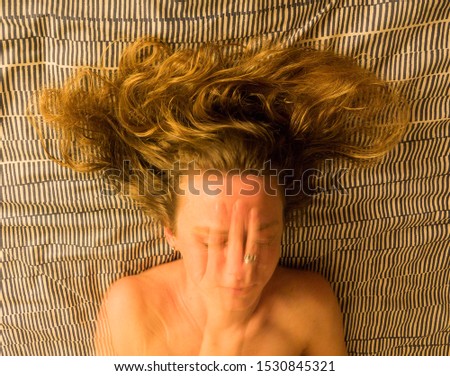 Colour close up of a woman's head and shoulders, lying on striped material, with her blonde hair spread out in a fan shape behind her, and her hand partially obscuring her face