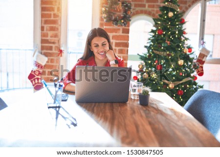 Beautiful woman sitting at the table working with laptop at home around christmas tree with a happy and cool smile on face. Lucky person.