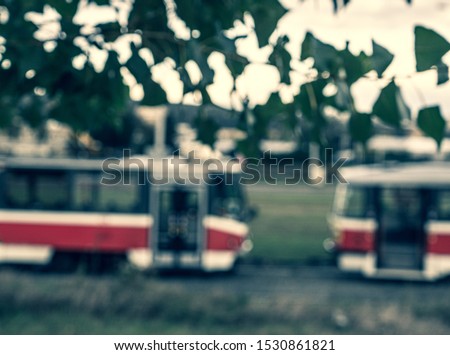 Blurry picture of two red tram carriages in Brno city with blurry leaves and intense sunlight in the top. Intentionally blurry transportation vehicles behind blurry green-white foreground elements.