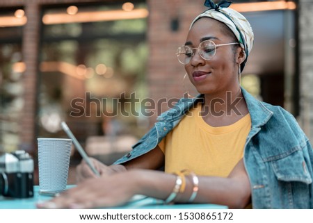 Cheerful trendy female is resting with coffee outdoors and writing in sketchbook on table. Website banner