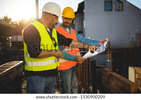 Men in hardhat and yellow and orange jacket posing on building site. stock photo