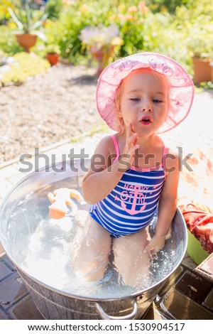 Adorable blonde baby girl 3 year old having bath at backyard. Little girl in a blue and white striped swimsuit and pink hat is in tin bathtub.
